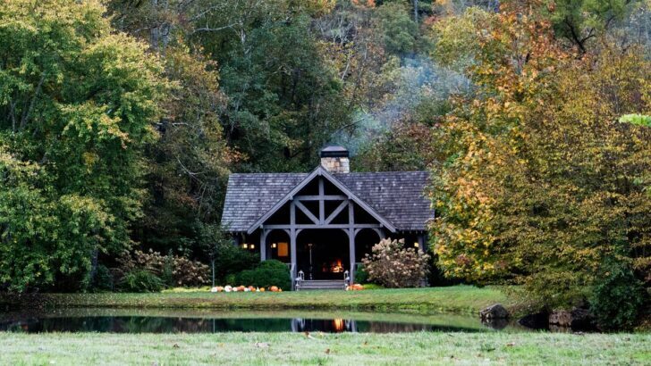 Cabaña rústica rodeada de árboles en un bosque otoñal con un pequeño lago en frente