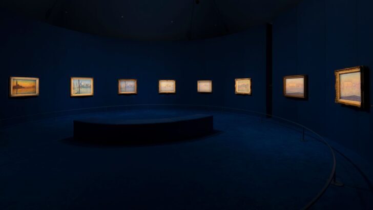 Sala de exposición con pinturas enmarcadas iluminadas en un fondo azul oscuro