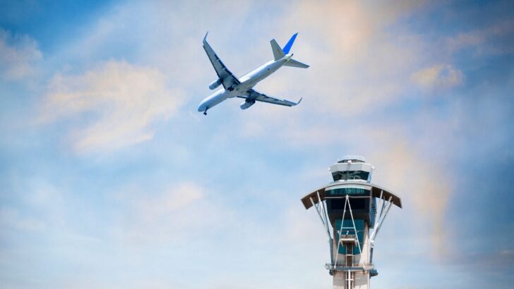 Avión volando cerca de una torre de control bajo un cielo azul con nubes