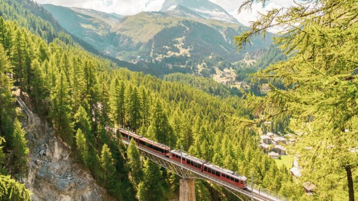Tren rojo cruzando un puente en un paisaje alpino con montañas y árboles